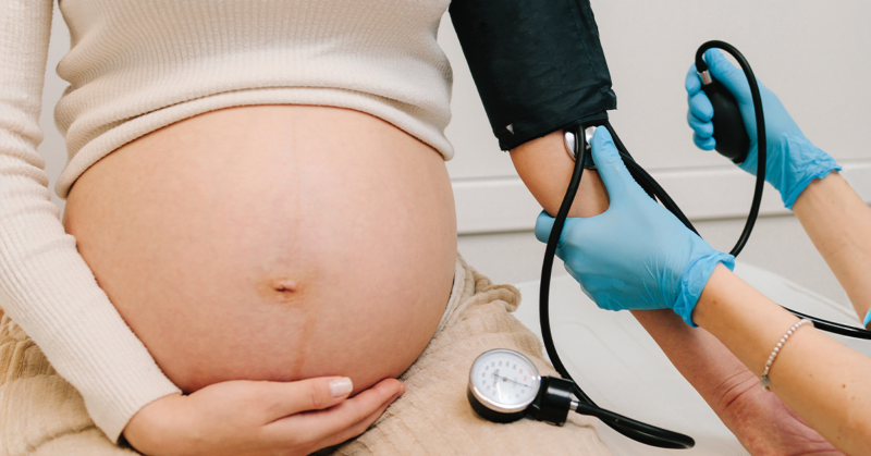 A pregnant woman wearing a white top holds her bare belly. A person wearing blue gloves checks her blood pressure, representing preeclampsia.