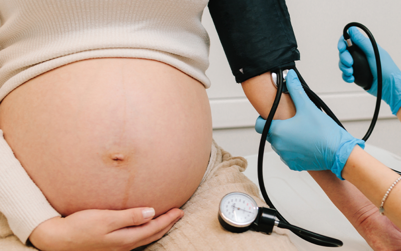 A pregnant woman wearing a white top holds her bare belly. A person wearing blue gloves checks her blood pressure, representing preeclampsia.