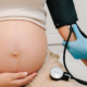 A pregnant woman wearing a white top holds her bare belly. A person wearing blue gloves checks her blood pressure, representing preeclampsia.