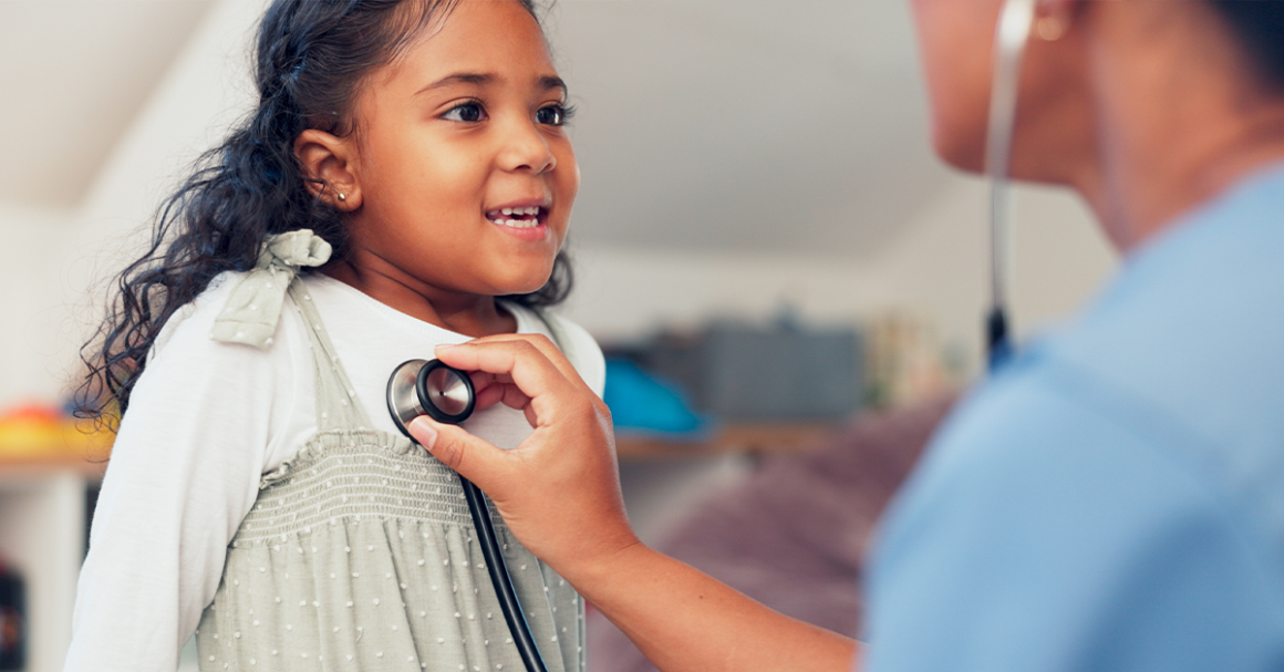 A young girl smiles as a healthcare professional listens to her heartbeat with a stethoscope.