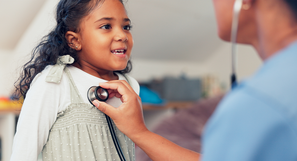 A young girl smiles as a healthcare professional listens to her heartbeat with a stethoscope.