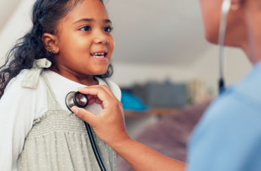 A young girl smiles as a healthcare professional listens to her heartbeat with a stethoscope.