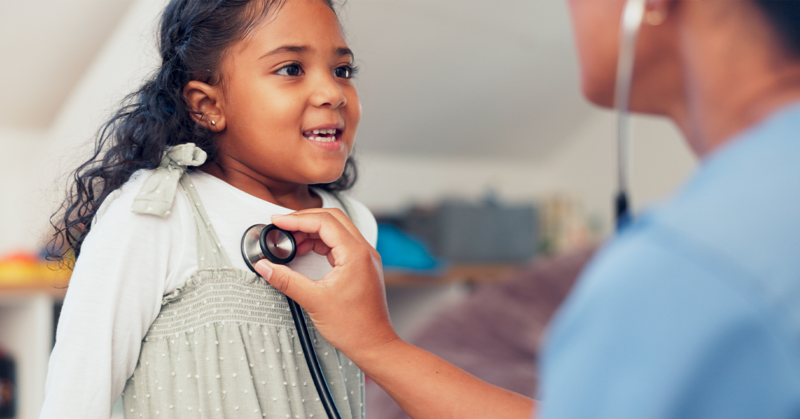 A young girl smiles as a healthcare professional listens to her heartbeat with a stethoscope.