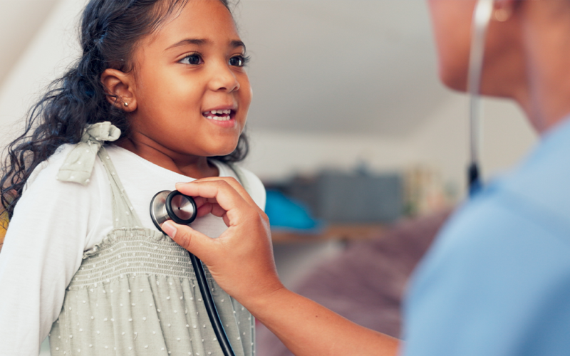 A young girl smiles as a healthcare professional listens to her heartbeat with a stethoscope.