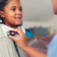 A young girl smiles as a healthcare professional listens to her heartbeat with a stethoscope.