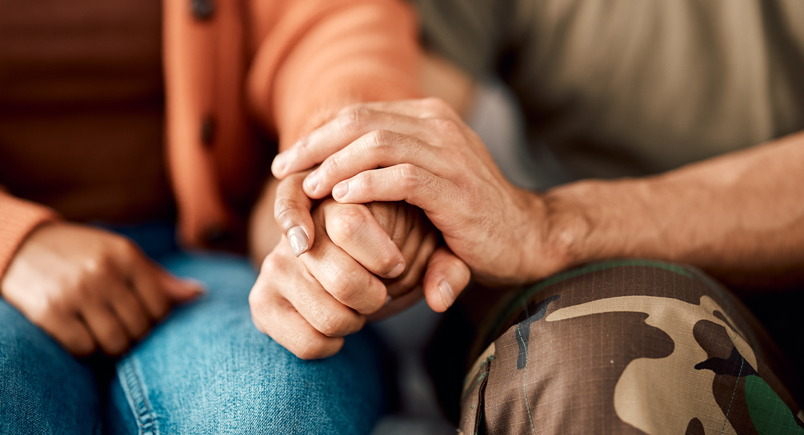 A close-up of two people sitting side by side, one in a brown sweater holding hands with another in camo pants.