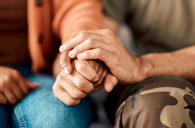A close-up of two people sitting side by side, one in a brown sweater holding hands with another in camo pants.
