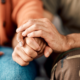 A close-up of two people sitting side by side, one in a brown sweater holding hands with another in camo pants.
