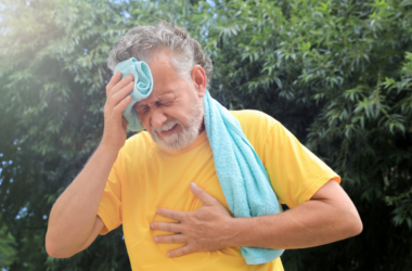Elderly man in a yellow shirt wipes sweat from his brow with a towel. He appears distressed, clutching his chest, against a backdrop of green trees.