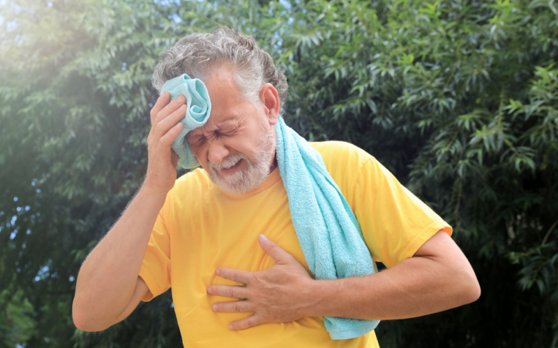 Elderly man in a yellow shirt wipes sweat from his brow with a towel. He appears distressed, clutching his chest, against a backdrop of green trees.