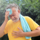 Elderly man in a yellow shirt wipes sweat from his brow with a towel. He appears distressed, clutching his chest, against a backdrop of green trees.