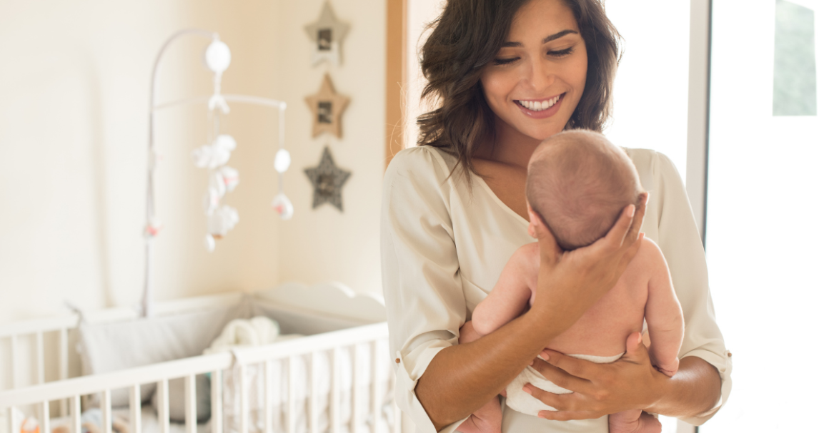 A smiling woman holds a baby in a cozy nursery with a crib and star decorations. The scene conveys warmth and tenderness in a bright setting, representing good mental health after giving birth.