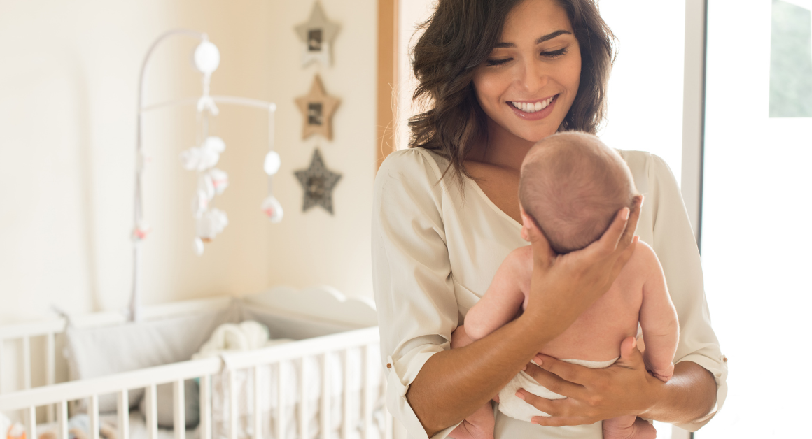 A smiling woman holds a baby in a cozy nursery with a crib and star decorations. The scene conveys warmth and tenderness in a bright setting, representing good mental health after giving birth.