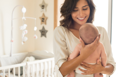A smiling woman holds a baby in a cozy nursery with a crib and star decorations. The scene conveys warmth and tenderness in a bright setting, representing good mental health after giving birth.