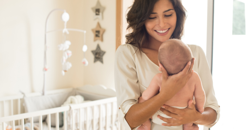 A smiling woman holds a baby in a cozy nursery with a crib and star decorations. The scene conveys warmth and tenderness in a bright setting, representing good mental health after giving birth.