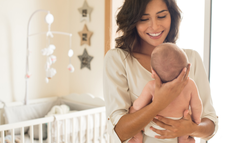 A smiling woman holds a baby in a cozy nursery with a crib and star decorations. The scene conveys warmth and tenderness in a bright setting, representing good mental health after giving birth.