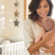 A smiling woman holds a baby in a cozy nursery with a crib and star decorations. The scene conveys warmth and tenderness in a bright setting, representing good mental health after giving birth.