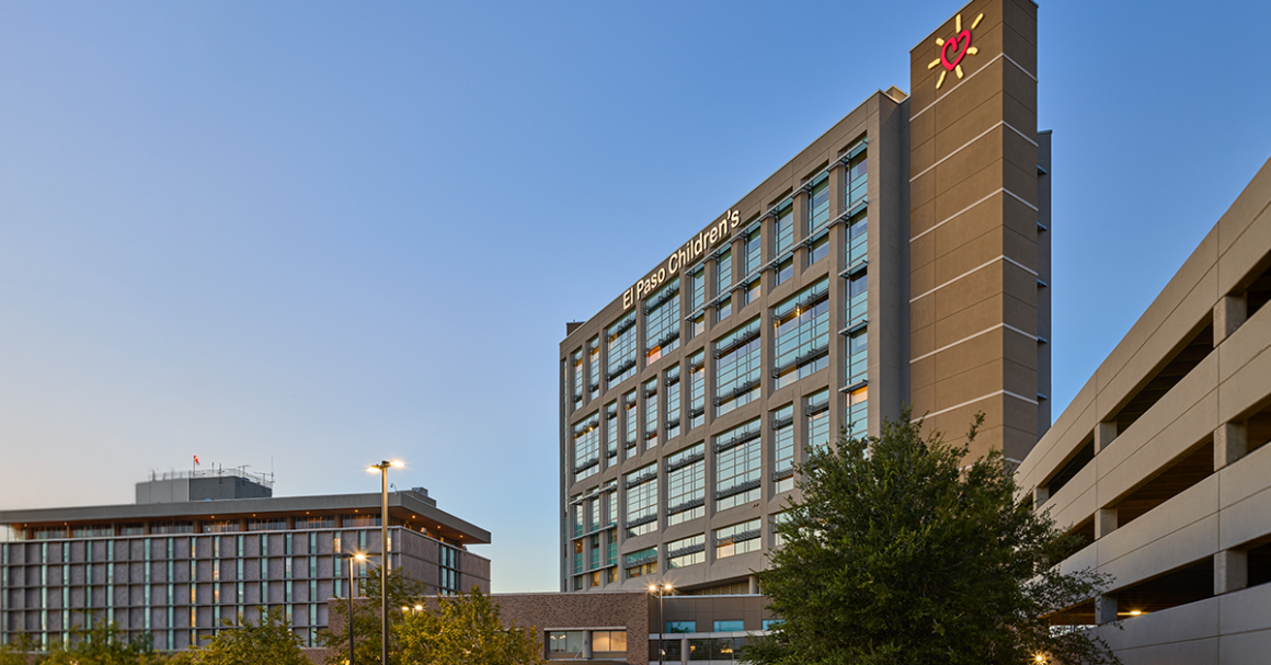 Modern hospital building with glass windows at dusk, labeled "El Paso Children's." Nearby, a tree and parking structure under a clear sky.