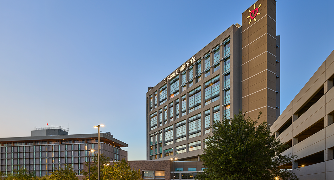 Modern hospital building with glass windows at dusk, labeled "El Paso Children's." Nearby, a tree and parking structure under a clear sky.