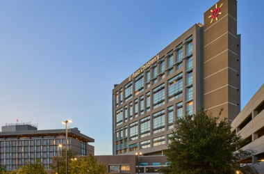 Modern hospital building with glass windows at dusk, labeled "El Paso Children's." Nearby, a tree and parking structure under a clear sky.