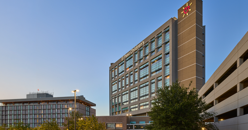 Modern hospital building with glass windows at dusk, labeled "El Paso Children's." Nearby, a tree and parking structure under a clear sky.