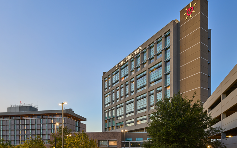 Modern hospital building with glass windows at dusk, labeled "El Paso Children's." Nearby, a tree and parking structure under a clear sky.