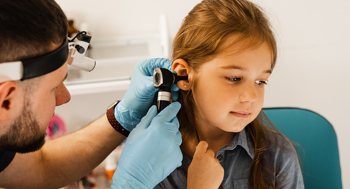 A healthcare professional examines a young girl's ear with an otoscope. The child looks calm and curious.