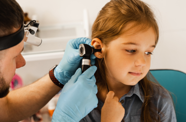 A healthcare professional examines a young girl's ear with an otoscope. The child looks calm and curious.