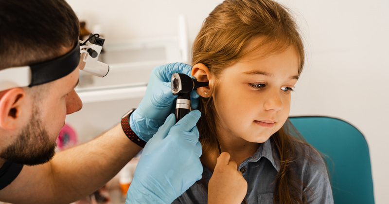 A healthcare professional examines a young girl's ear with an otoscope. The child looks calm and curious.