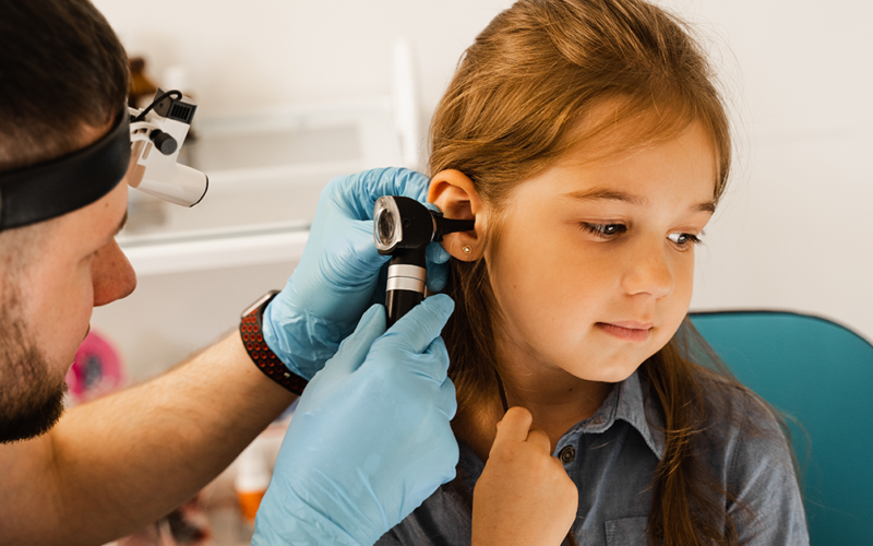 A healthcare professional examines a young girl's ear with an otoscope. The child looks calm and curious.