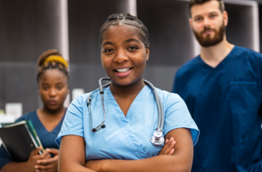 A confident young nurse with a stethoscope smiles in the foreground. Behind her, two medical professionals stand, conveying teamwork in a healthcare setting.