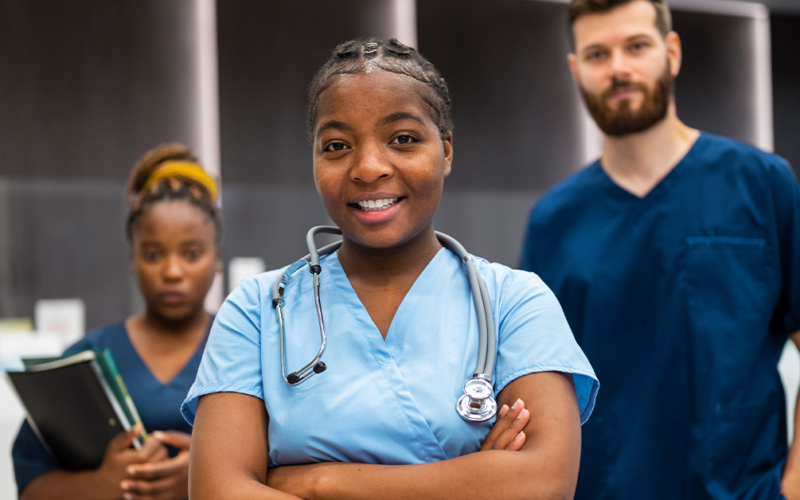 A confident young nurse with a stethoscope smiles in the foreground. Behind her, two medical professionals stand, conveying teamwork in a healthcare setting.