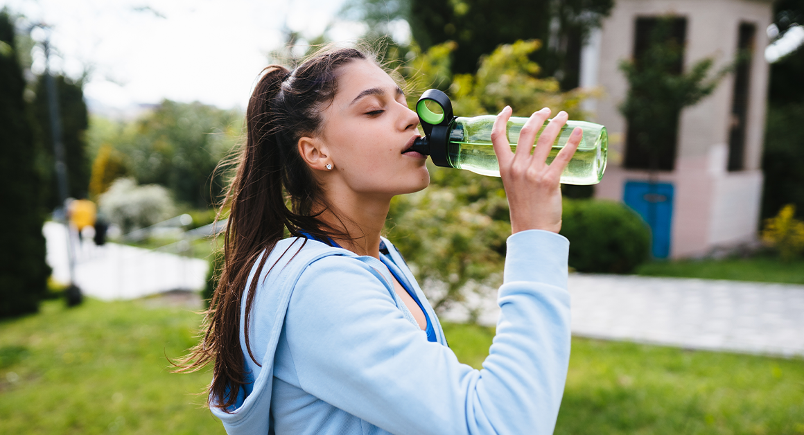 Woman in a blue hoodie drinks from a green water bottle outdoors. She's in a lush park setting, taking a refreshing break, conveying relaxation.
