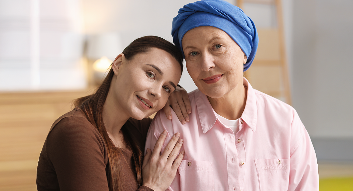 A loving portrait of two women; one with a blue headscarf, the other resting her head on her shoulder. They are indoors, representing National Cancer Survivors Day.