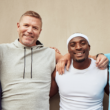 Four men smiling and standing arm-in-arm against a beige wall. They wear casual sports attire, exuding camaraderie, diversity, and happiness, representing Men's Health Month.