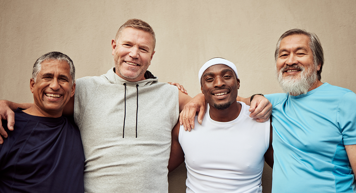 Four men smiling and standing arm-in-arm against a beige wall. They wear casual sports attire, exuding camaraderie, diversity, and happiness, representing Men's Health Month.