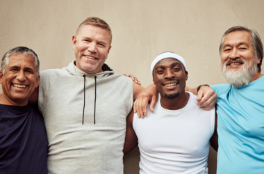 Four men smiling and standing arm-in-arm against a beige wall. They wear casual sports attire, exuding camaraderie, diversity, and happiness, representing Men's Health Month.