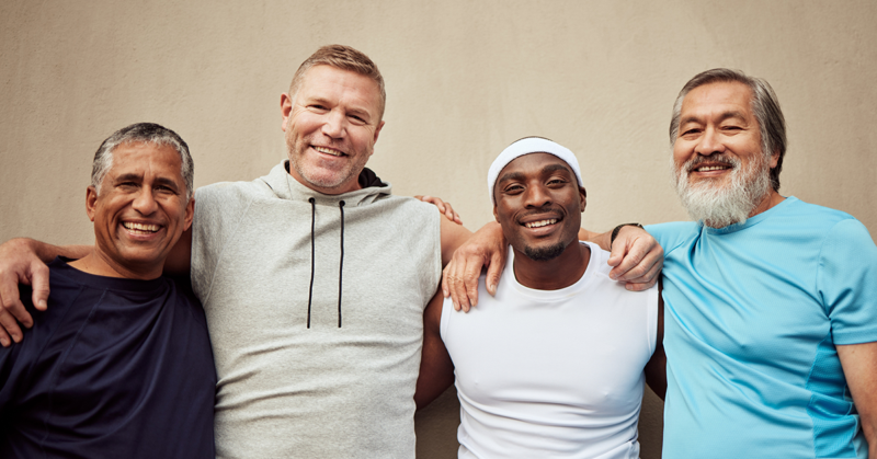Four men smiling and standing arm-in-arm against a beige wall. They wear casual sports attire, exuding camaraderie, diversity, and happiness, representing Men's Health Month.