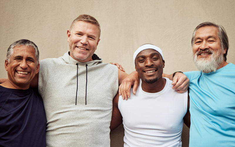 Four men smiling and standing arm-in-arm against a beige wall. They wear casual sports attire, exuding camaraderie, diversity, and happiness, representing Men's Health Month.