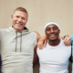 Four men smiling and standing arm-in-arm against a beige wall. They wear casual sports attire, exuding camaraderie, diversity, and happiness, representing Men's Health Month.