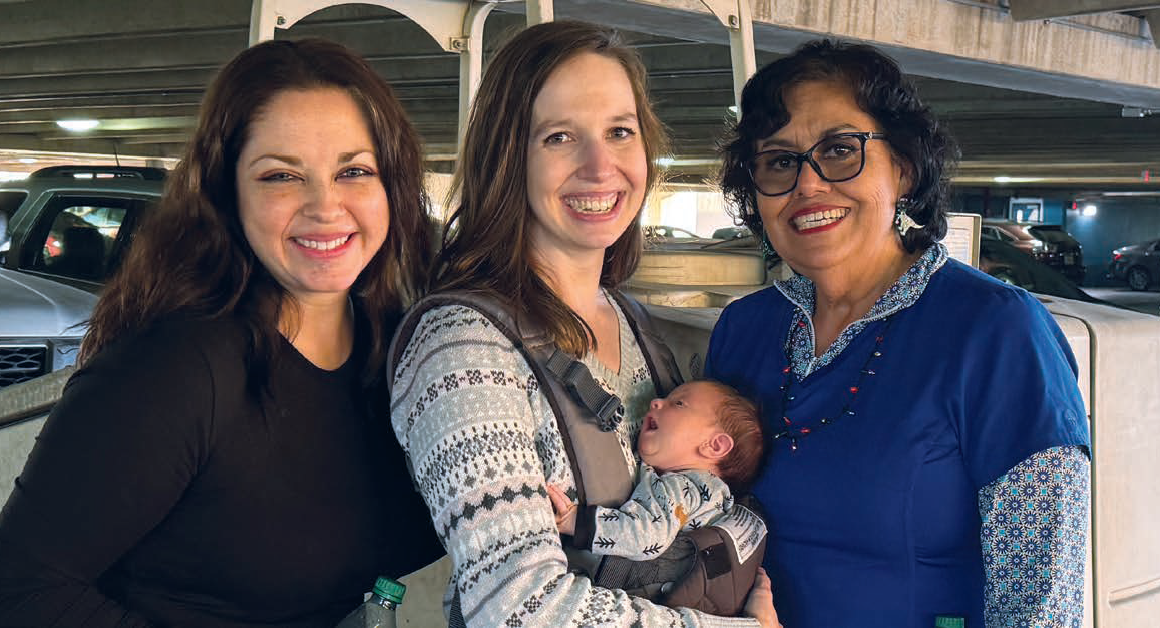 Three adults smile together while one holds a baby in a front carrier.