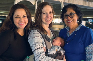 Three adults smile together while one holds a baby in a front carrier.