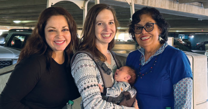 Three adults smile together while one holds a baby in a front carrier.
