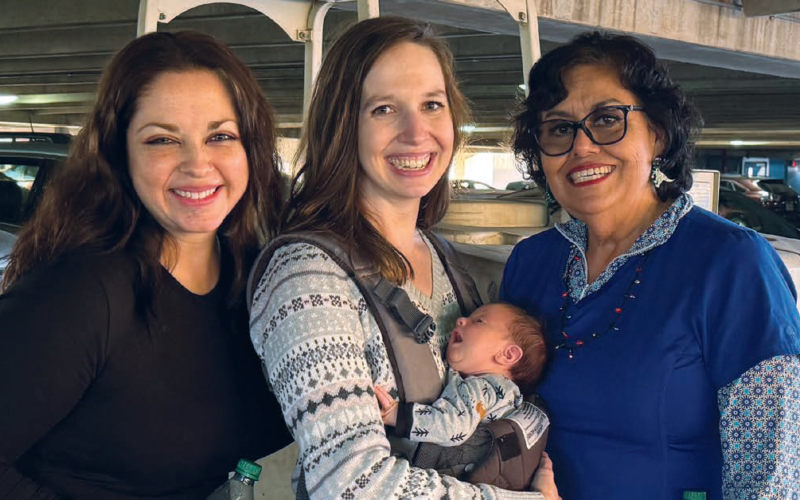 Three adults smile together while one holds a baby in a front carrier.