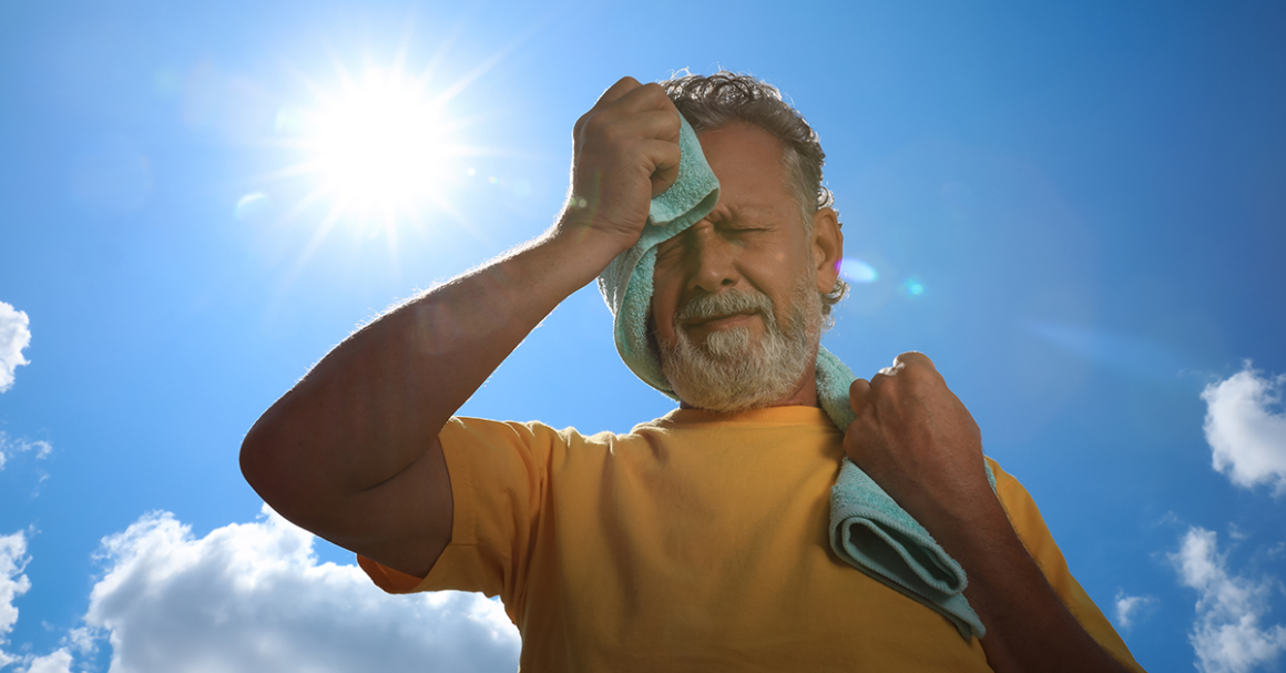 Man wipes sweat from his face with a towel while standing outdoors under the hot sun.