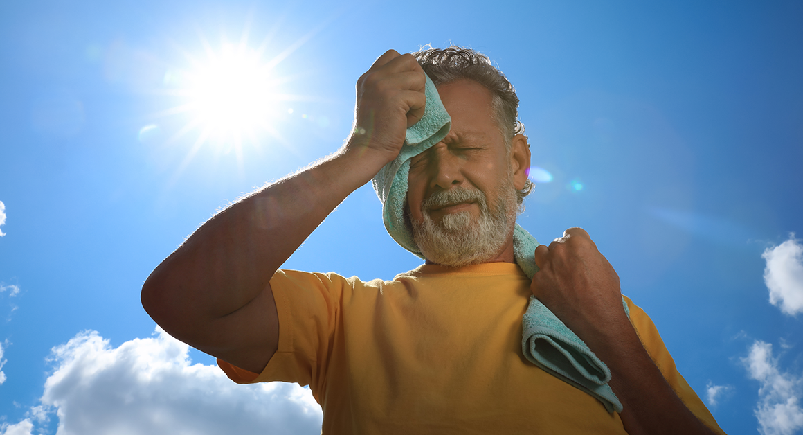 Man wipes sweat from his face with a towel while standing outdoors under the hot sun.