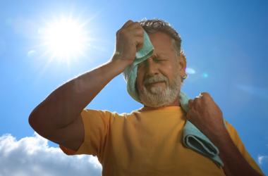 Man wipes sweat from his face with a towel while standing outdoors under the hot sun.