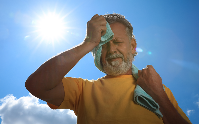Man wipes sweat from his face with a towel while standing outdoors under the hot sun.
