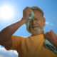 Man wipes sweat from his face with a towel while standing outdoors under the hot sun.