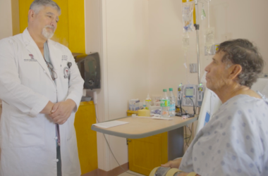 Dr. Philip E. Fiddler, Director of the UMC Burn Center, talks to Javier Diaz, a patient sitting in a hospital bed. The room has medical equipment.